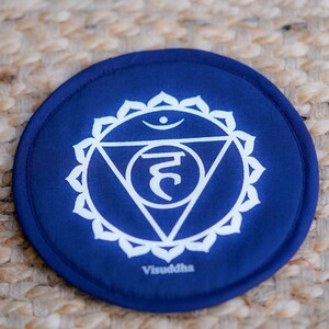 Op de afbeelding: Een blauw rond kussen met een wit symbool en het woord "Visuddha" erop gedrukt. Het symbool is een driehoek met een cirkel bovenaan en een halvemaanvorm in de driehoek. Het symbool wordt omringd door een cirkel met 10 bloemblaadjes.