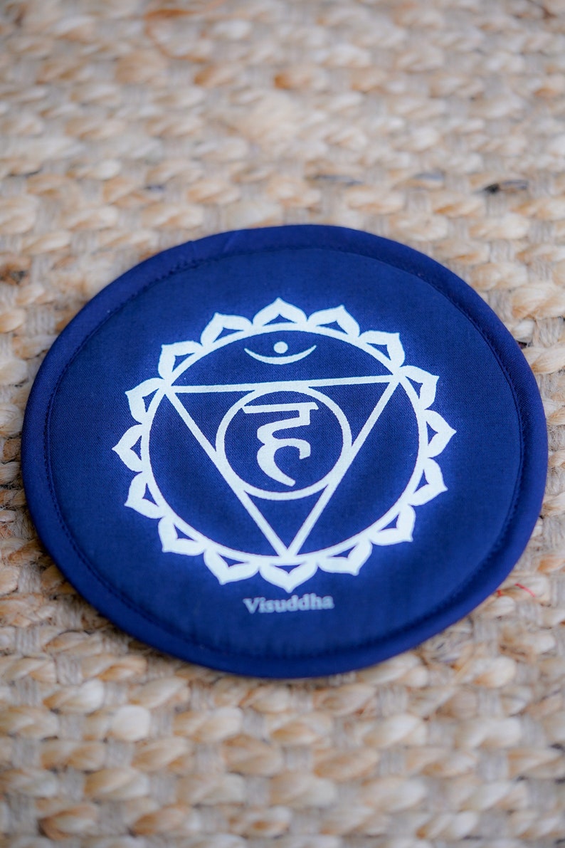 Op de afbeelding: Een blauw rond kussen met een wit symbool en het woord "Visuddha" erop gedrukt. Het symbool is een driehoek met een cirkel bovenaan en een halvemaanvorm in de driehoek. Het symbool wordt omringd door een cirkel met 10 bloemblaadjes.
