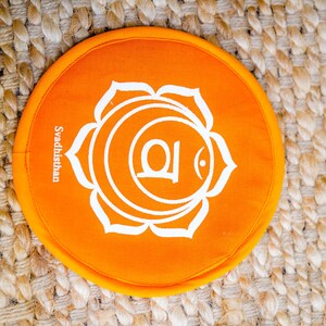 Op de afbeelding: Rond oranje kussen met een wit lotusbloemontwerp en het woord "Svadhisthan" erop gedrukt.