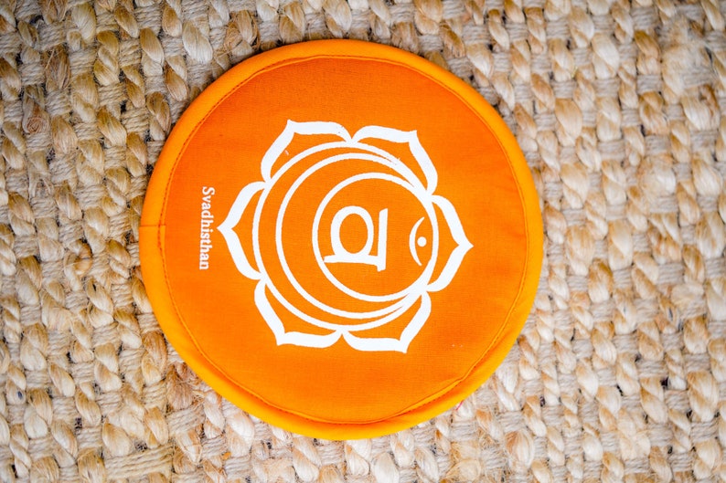 Op de afbeelding: Rond oranje kussen met een wit lotusbloemontwerp en het woord "Svadhisthan" erop gedrukt.