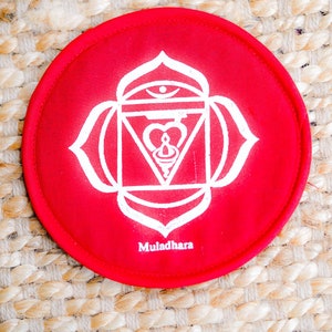 Op de afbeelding: Rond kussen van rode stof met een wit mandala-ontwerp en het woord "Muladhara" eronder gedrukt. Het kussen ligt op een geweven strooppervlak.