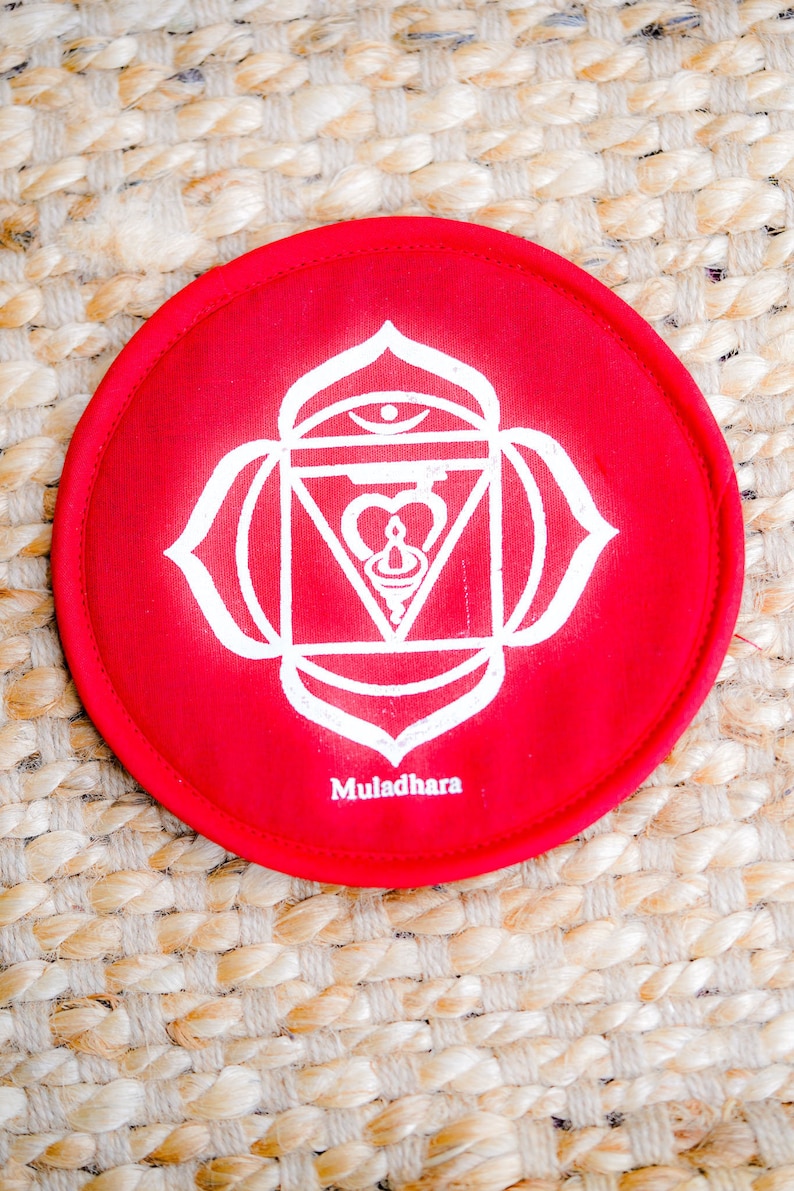 Op de afbeelding: Rond kussen van rode stof met een wit mandala-ontwerp en het woord "Muladhara" eronder gedrukt. Het kussen ligt op een geweven strooppervlak.
