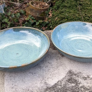 May include: Two blue ceramic bowls with a light blue glaze and a brown rim. The bowls are sitting on a gray concrete surface.