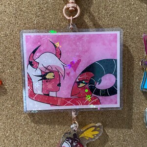 May include: A pink and red glitter keychain featuring a cartoon couple with hearts and stars. The keychain has a clear acrylic charm with a pink and white design.