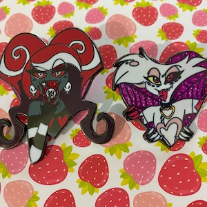 Könnte beinhalten: Zwei Emaille-Pins mit Charakteren aus der Zeichentrickserie Helluva Boss. Der erste Pin ist rot und schwarz und zeigt eine weibliche Figur mit roten Haaren und Hörnern. Der zweite Pin ist lila und weiß und zeigt eine weibliche Figur mit weißem Fell und Hörnern. Beide Pins sind herzförmig.