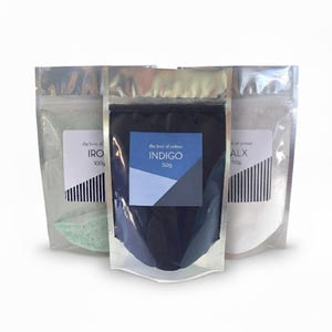 Peut inclure: Trois sachets en aluminium contenant des poudres colorées. Le sachet central est indigo, avec une étiquette indiquant "INDIGO 50g". Les autres sachets contiennent IRO 100g et ALX 150g. Chaque sachet porte l'inscription "the love of colour".