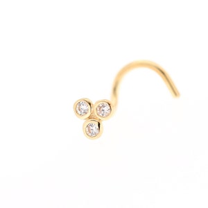 Könnte beinhalten: Goldener Nasenring mit drei kleinen, klaren Zirkonia-Steinen, die in einer dreieckigen Form gefasst sind.
