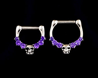 Puede incluir: Dos anillos de septum plateados con un motivo de calavera y gemas moradas. Los anillos tienen un diseño de bisagra. Las gemas están dispuestas en forma de arco. El fondo es negro.