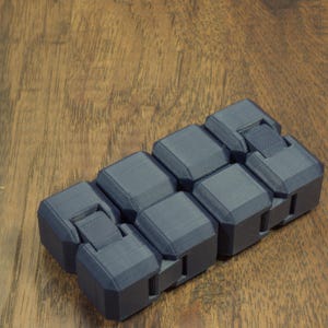 May include: A dark gray fidget toy with a segmented design. The toy is made of several connected blocks that can be moved and manipulated. The blocks have a textured surface and are arranged in a rectangular shape. The toy is on a wooden surface.