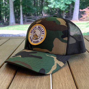 May include: A camouflage trucker hat with a black mesh back. The hat has a yellow and purple patch with the text "Death Valley Baton Rouge, LA".
