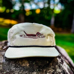 May include: A beige, flat-brimmed cap with a black rope detail and a brown embroidered dachshund dog in water. The dog is holding a stick. The cap is sitting on a large, textured rock.