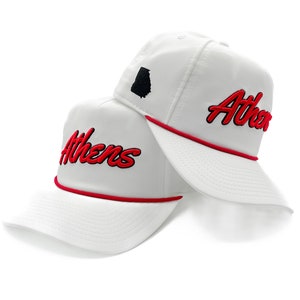 May include: Two white baseball caps featuring a red rope trim across the base of the crown. Each cap displays the word Athens embroidered in red script with a black outline. One cap includes a small black silhouette of the state of Georgia.