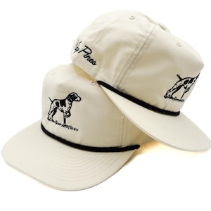 May include: Two cream-colored baseball caps with black rope trim and a flat brim. Each cap features a black and white dog graphic and the text "Lily Pines" embroidered in black. The caps are stacked, showcasing the design.