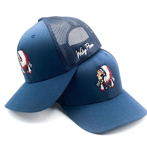May include: Two navy blue trucker hats stacked on top of each other. Each hat features a mesh back and a curved brim. The front of each hat displays an embroidered Native American chief design in red, white, and yellow. The text "Wsky Pines" is written on the top hat.