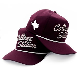 May include: Two maroon baseball caps with white stitching. The front of the caps feature the text "College Station" in a white cursive font. The top of one cap has a white outline of the state of Texas.