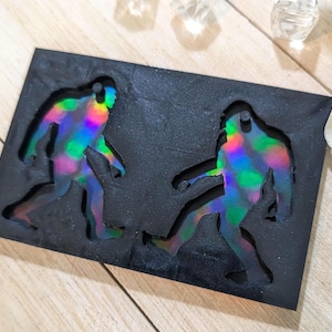 May include: A black rectangular mold featuring two Bigfoot figures with iridescent, rainbow-colored details. The figures are in a walking pose, with one leg forward. The mold is on a light-colored wooden surface.