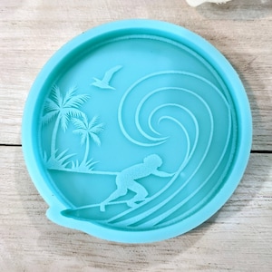 May include: A light blue silicone mold with a design of a surfer riding a wave, palm trees, and a bird. The mold is round and has a smooth surface.