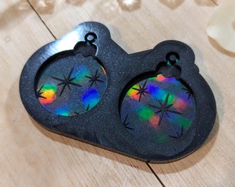 May include: A black resin mold with two circular cavities, each featuring a holographic, iridescent design of a Christmas ornament with starbursts. The mold is designed for crafting holiday decorations or ornaments.