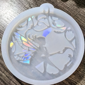 May include: A round, white silicone mold with a fairy design. The mold features a fairy with iridescent wings and a detailed face. The mold has a small loop at the top for hanging. The design is perfect for resin art projects.