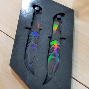 May include: Two decorative knives with a Scream mask design, presented in a black, glittery case. The blades have a rainbow iridescent finish, adding a striking visual element. The knives are designed with a serrated edge.