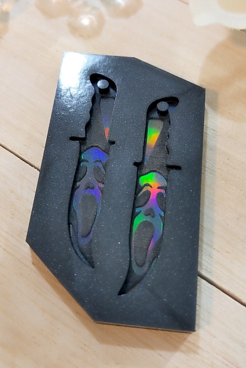 May include: Two decorative knives with a Scream mask design, presented in a black, glittery case. The blades have a rainbow iridescent finish, adding a striking visual element. The knives are designed with a serrated edge.