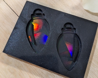 May include: Two iridescent Christmas ornament-shaped pendants are displayed in a black, textured box. The pendants have a rainbow effect, reflecting various colors. The box is set on a light-colored wooden surface.