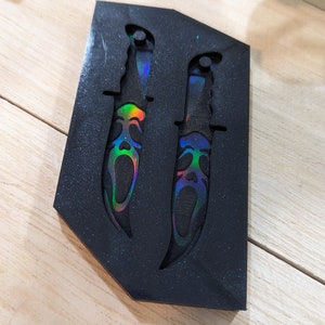 May include: Two black decorative knives with iridescent blades and handles, set in a black foam mold. The blades feature a Scream mask design with rainbow-colored accents. The knives are displayed on a light wood surface.