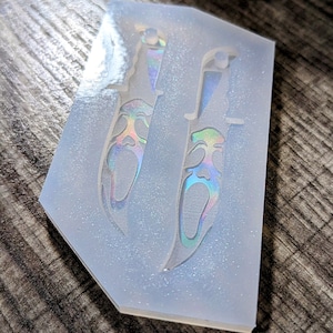 May include: A clear silicone mold with two knife-shaped cavities. The knife designs feature a face with a screaming expression, with iridescent coloring. The mold is on a wooden surface.