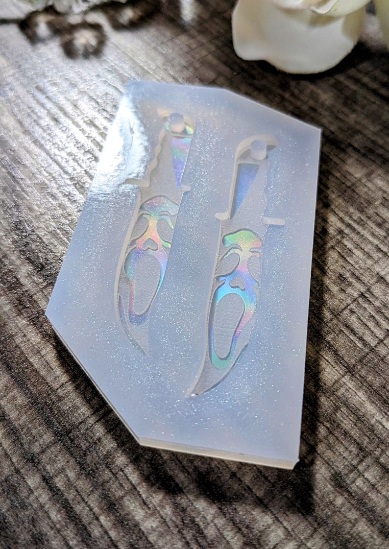 May include: A clear silicone mold with two knife-shaped cavities. The knife designs feature a face with a screaming expression, with iridescent coloring. The mold is on a wooden surface.