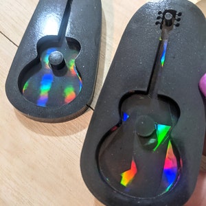 May include: Two black guitar-shaped molds with a holographic, rainbow-colored design inside. The molds are oval-shaped and sit on a light-colored wooden surface. The guitar shape is clearly defined within the mold.