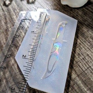May include: A clear silicone mold with a knife design, approximately 5 inches long. The knife has a decorative handle and a holographic effect. A ruler is placed next to the mold for measurement. The mold is on a wooden surface.