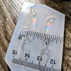 May include: A clear silicone mold with two dagger shapes, each approximately 1.25 inches long. The daggers have decorative handles and iridescent accents. A ruler is placed next to the mold for measurement.