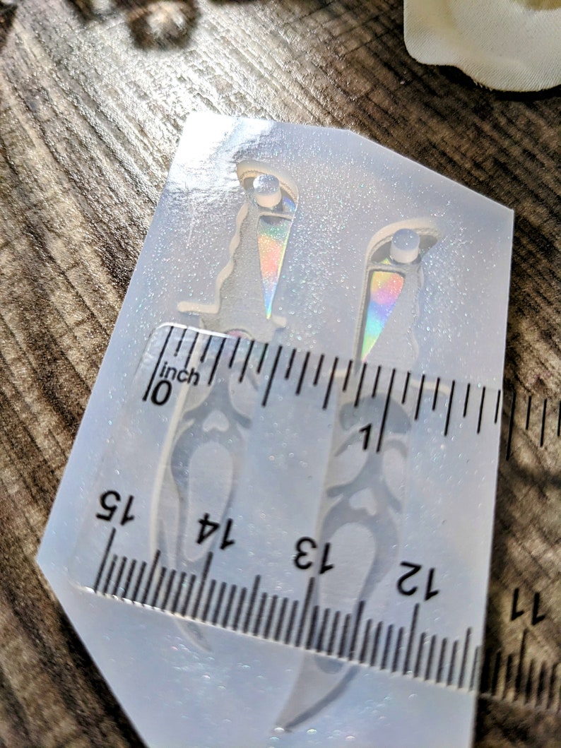 May include: A clear silicone mold with two dagger shapes, each approximately 1.25 inches long. The daggers have decorative handles and iridescent accents. A ruler is placed next to the mold for measurement.