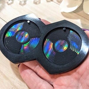 May include: Two black resin coasters with a holographic vinyl record design. The coasters are shaped like a figure eight.