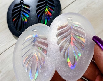 May include: Two leaf-shaped molds, one black and one clear, with a leaf design. The black mold has a holographic effect. The clear mold has a leaf design with a holographic effect. The molds are on a light-colored surface.