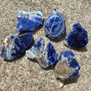 May include: Six rough, natural, blue and white, sodalite stones. The stones are various sizes and shapes, with some showing white and brown veins.