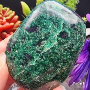 May include: A polished, oval-shaped gemstone with a vibrant green hue, speckled with darker green and small flecks of deep red. The stone has a smooth, glossy surface and a natural crack running across it. The background features colorful plants.