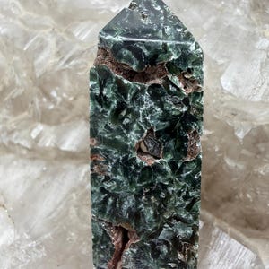 May include: A polished seraphinite crystal tower with a pointed top, featuring a deep forest green color with iridescent, feathery patterns and visible natural brown matrix inclusions throughout the textured, crystalline surface.