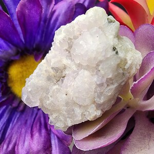 May include: A close-up of a cluster of light-colored quartz crystals. The crystals are resting on a bed of purple and lavender artificial flowers with yellow centers and red and yellow accents.