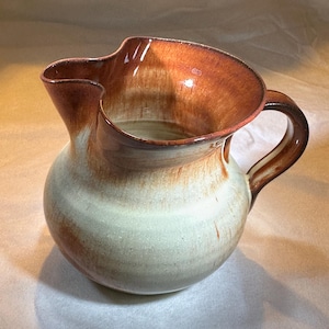 May include: A ceramic pitcher with a rounded body and a handle. The pitcher has a light beige base that fades into a brown rim and handle. The spout is designed for easy pouring. This decorative pitcher is suitable for serving beverages.