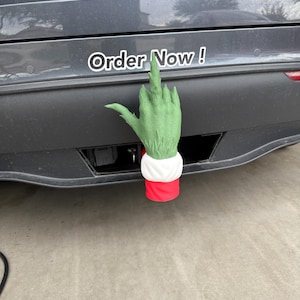 May include: A green hand with a white cuff and red sleeve, attached to a car's bumper. The hand is giving the middle finger. Above the hand is a white sticker with the words "Order Now!"