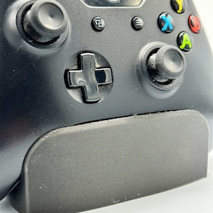 May include: A black Xbox controller with a black charging stand. The controller has a black d-pad, black buttons, and a black Xbox logo.