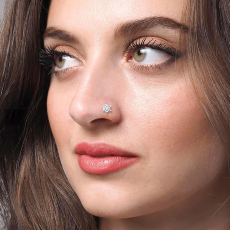 May include: A close-up of a person's face with a small, silver, flower-shaped nose ring with clear stones.