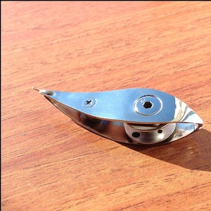 Könnte beinhalten: Ein silberner Segelboot-Klampen aus Metall mit einem kleinen Loch in der Mitte. Der Klampen befindet sich auf einer Holzoberfläche.