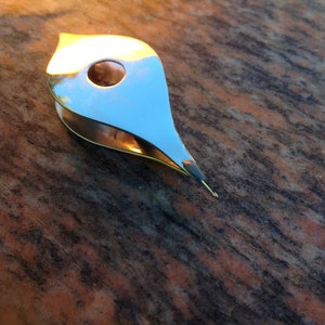 May include: A silver metal tool with a teardrop shape and a small hole in the center. The tool is designed for crafting and is lying on a brown and black textured surface.