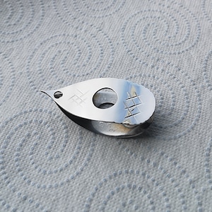 May include: A silver metal tool shaped like a teardrop with a small hole in the centre. The tool has a textured surface with a grid pattern.