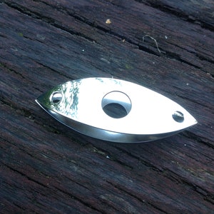 Op de afbeelding: Een gepolijste zilverkleurige metalen sigarenknipper met een langwerpige, oogvormige vorm. Het gereedschap heeft een grote ronde opening in het midden om te knippen en twee kleinere ronde gaten aan de spitse uiteinden, met een glad oppervlak.