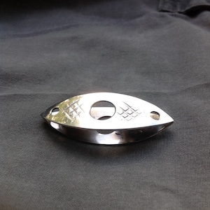 Op de afbeelding: Een zilverkleurig metalen frivolité-schuitje met een langwerpig, oogvormig profiel. Het oppervlak is voorzien van een centrale ronde uitsparing en twee kleinere ronde gaten aan de spitse uiteinden, geaccentueerd door twee kleine rasterpatronen.
