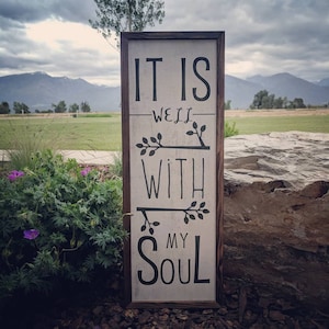 May include: A rustic wooden sign with a white background and black lettering. The sign reads "It is well with my soul". The sign is decorated with small black branches with leaves.