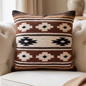 May include: A square throw pillow featuring a woven pattern of horizontal stripes in shades of brown, cream, and black. The design includes repeating geometric diamond motifs across the center and top sections, with a textured fabric finish.